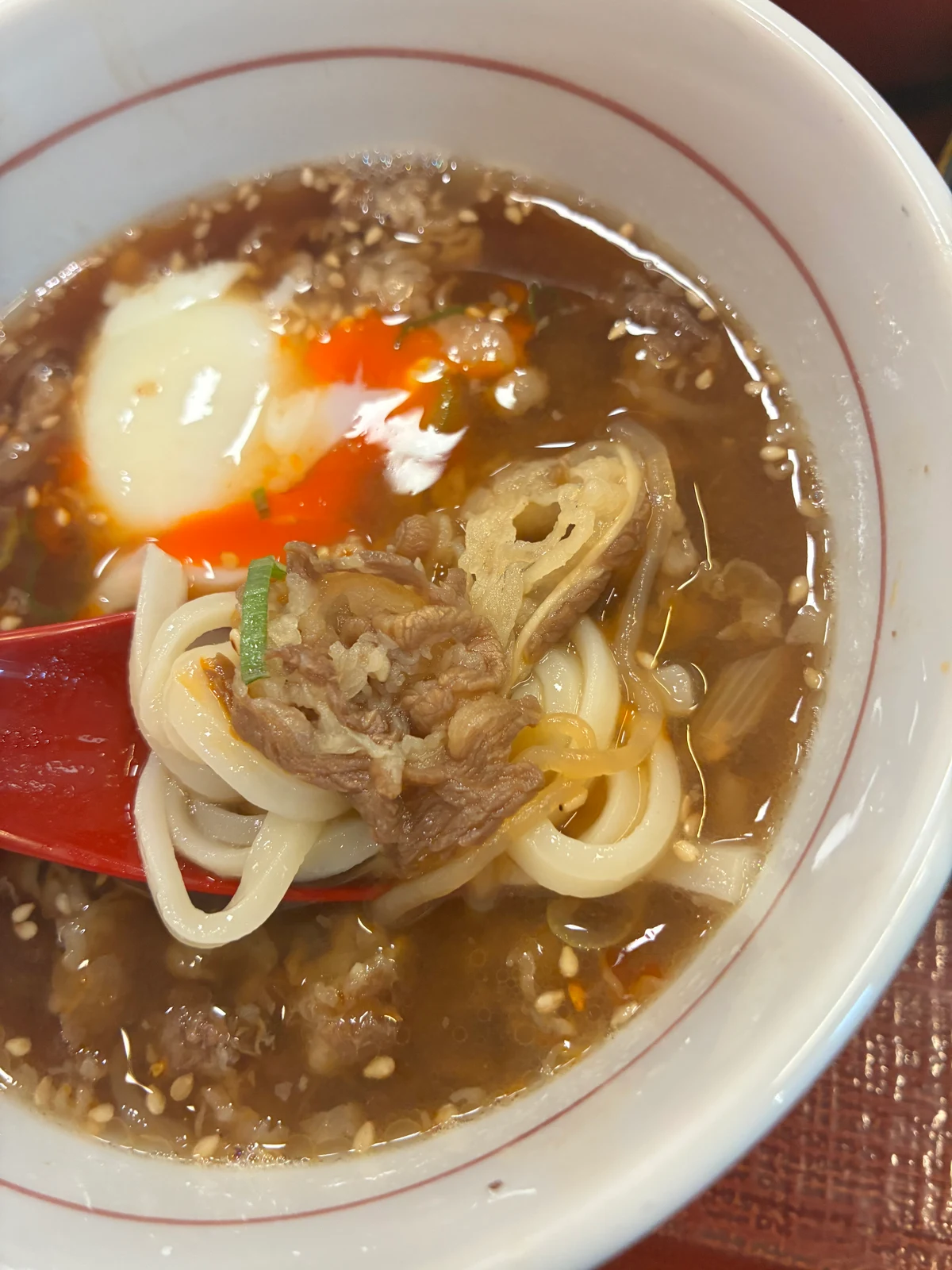 れんげにのせたうどんと薄切り牛肉、背後の温たま入りつけ汁